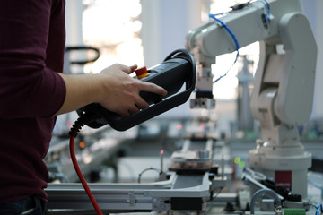 Close up of man's hand holding teach panel  to control robot arm which is integrated on smart factory production line. industry 4.0 automation line equipped with sensors and robot at background.