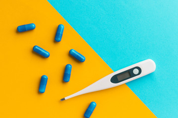 Digital thermometer and blue pills isolated on bright blue-yellow background. Epidemic, painkillers, healthcare and treatment concept. Flat lay. Top view with copy space.