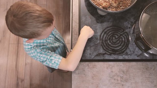 Child reaches for hot electric stove and play on kitchen. Kid risking accident. Dangerous situation at home. Concept of burn children. Toddler touches pot of boiling water with his hand