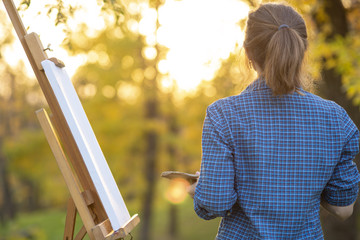 young woman artist drawing a picture on canvas on an easel in nature, a girl with a brush and a...