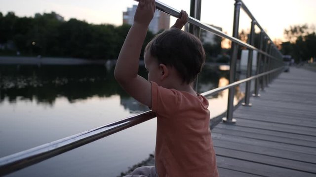Little boy sits on a wooden plank and looks on a water. Kid waves his hand