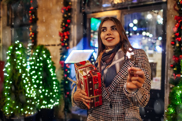 Obraz na płótnie Canvas Christmas, New Year concept. Woman burning sparkler on city street by decorations. Stylish girl holding gift box present