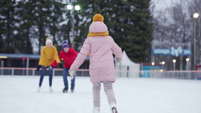 Follow rear view shot of adorable little girl learning to ice skate on outdoor skating rink. Cute kid skating in slow motion into her loving parents embrace