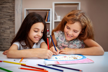 two beautiful teenage girls, a brunette and a blonde laugh, draw with colored pencils in an album on a white table, in the background are white shelves with books and toys