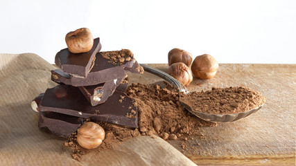 Broken dark chocolate bars, cocoa powder and hazelnuts on beige paper surface