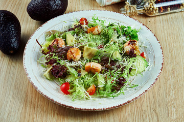 Tasty, fresh green salad with shrimp, lettuce, avocado, parmesan and cherry tomatoes on a white plate on a wooden background. Healthy nutrition. Close up on food.