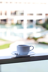 White mug on a white saucer with coffee or tea on the background of the outdoor pool on site.