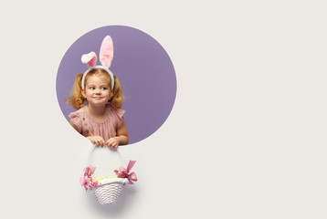 Obraz na płótnie Canvas Easter card. Cute little child girl with bunny ears holding basket of Easter eggs. Child in a round hole circle in colored purple and white background. Mockup and copy space for text