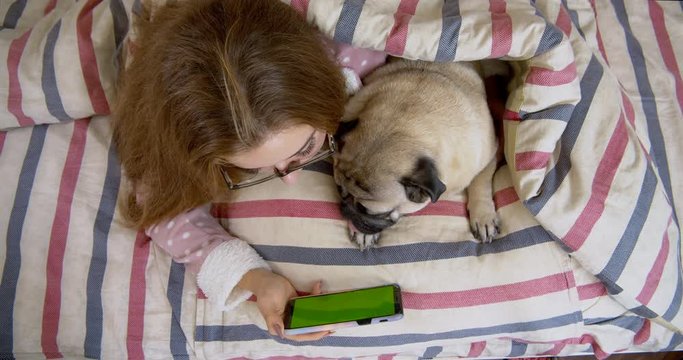 Beauty teen girl and her cute pug dog lying in the bed, watching, looking at smartphone. Green screen. Friendship with pet concept. Together forever, inseparable friends