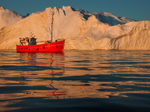 Red boat sailing past iceberg at sunset in Greenland, copy space