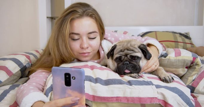 Beauty teen girl and her cute pug dog lying in the bed, taking selfie on smartphone. Friendship with pet concept. Together forever, inseparable friends