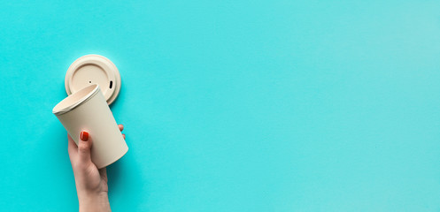 Stylish reusable eco coffee travel mug, bamboo cup with lid in hands, copy-space. Blue mint paper background, panoramic image. Minimalistic panoramic zero waste eco friendly lifestyle trend.