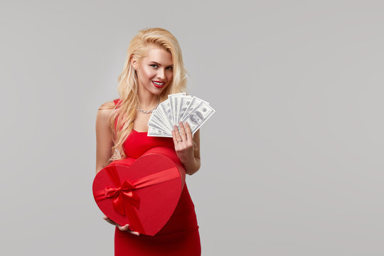 A young woman in a red dress holds in her hands cash banknotes money, dollars and a heart shaped gift box. Valentines Day and March 8