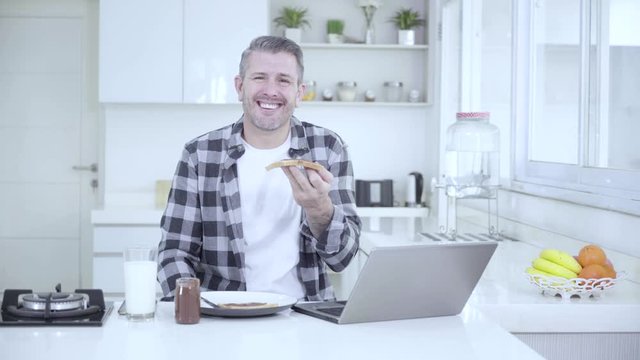 Funny Caucasian man laughing in the kitchen while holding a bread with laptop on the table. Shot in 4k resolution