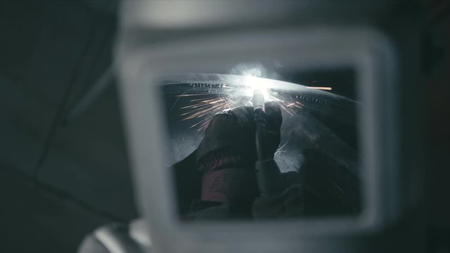 Forge workshop. Smithy. Worker in a welding hood helmet welds a part by electric welding. Sparks are reflected in the protective screen. Blacksmith makes iron product for manufacture. Slow motion.