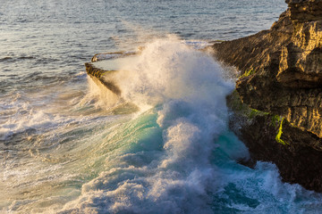 Giant wave crashing against rocky outcrop at Devil's Tear, on the coast of Nusa Lembongan Island, Bali, Indonesia. Tide pool visible behind wave on top of shore; ocean in the background. 