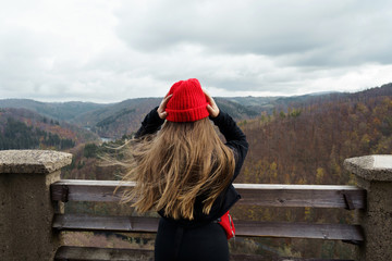 Fototapeta na wymiar Long-haired girl in a black jacket, a red knitted hat standing on top of a high tower in the mountains. Wind in the hair. Weekend and school travel concept. Faceless