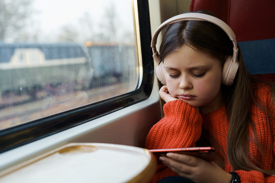 Teenager sad girl 12 years old in red jacket with headphones on her head sitting in suburban train watching a video on a mobile phone. Gadget addiction concept. Spring vacation. Daily trips to school