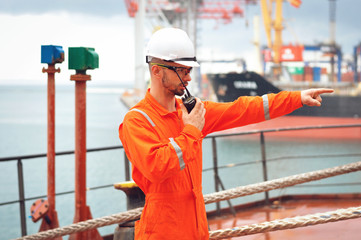 An experienced sailor in orange overalls and a white helmet speaks on a walkie-talkie and gives instructions to other workers.