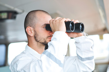 Sailor navigator, the third assistant to the captain looks through binoculars in a white overalls on the captain’s bridge. - 311107681