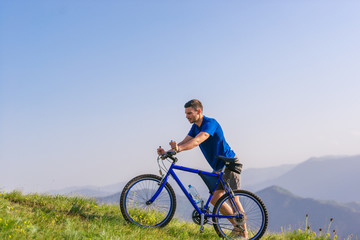 Fototapeta premium Tired fit mountain biker pushing his bike uphill at the top of the mountain on a sunny day with amazing view on a blue river.