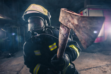 Portrait of a female firefighter while holding an axe and wearing an oxygen mask indoors surrounded...