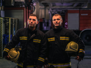 Portrait of two young firemen in uniform standing inside the fire station .