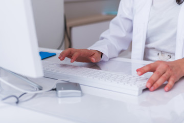 Close up hands of a female physician ( nurse ) typing on her white keyboard while sitting on a white desk in a hospital lobby.