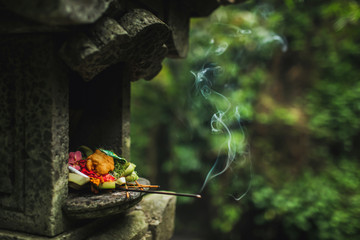 Traditional balinese Canang Sari offerings to gods and spirits with flowers, food and smoky aromatic sticks on dark green background. Indonesian culture and religion. Bali authentic travel concept.