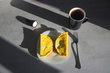 two semicircular omelettes on toasts on a white plate with a black cup of coffee and a black cone with dessert with beautiful long shadows on a gray kitchen table top view