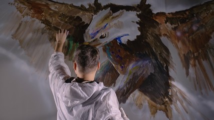 Artist designer draws an eagle on the wall. Craftsman decorator paints a picture with acrylic oil color. Painter painter dressed in a paint coat. Indoor. Dark magic cinematic look.