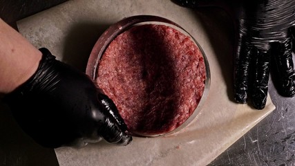 Cook makes meat medallions for burgers. Chef in black food gloves makes cutlet. Cutlets are leveled in steel ring in an even medallion. On top of pouring spices pepper and salt.