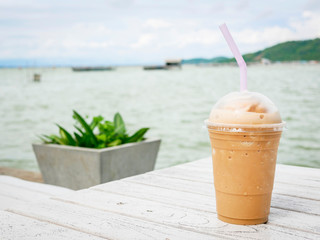 Iced coffee in a plastic cup on the white wooden table. Sea and mountain backdrop.  Y