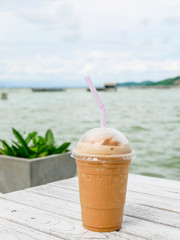Iced coffee in a plastic cup on the white wooden table. Sea and mountain backdrop.  Y