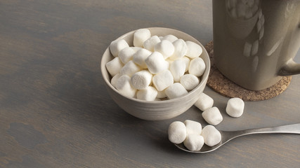 Small white marshmallow in a cream colour bowl, accompanied by a cup of hot beverage.