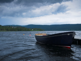 Naklejka premium Dark row boat tied to a dock on beautiful lake Hurdalssjoen in Norway, with storm clouds overhead. Beautiful Europe Travel Scene.