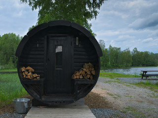 Round wooden outdoor sauna with stacks of wood for heating, by a lake with nearby trees and a picnic table.