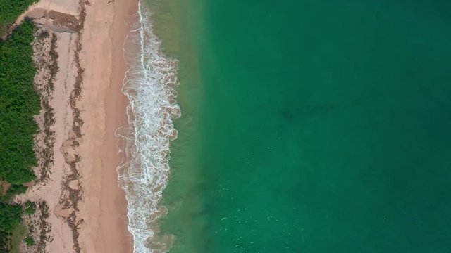 Aerial bird's eye view of ocean waves crashing against an empty beach. Big blue powerful waves rolling on tropical pink sand beach from above. Sunny day over the sea. Huge swell hitting shoreline.