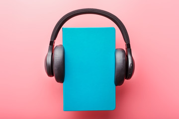 Headphones are worn on a book in a blue hardcover on a pink background, top view.