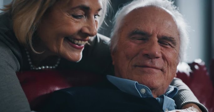 Close up of a lovely senior woman is hugging with affection her husband as a sign of forever love and respect to each other in on sofa in living room at home.