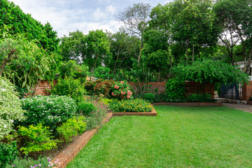 English cottage garden, colorful flowering plant on smooth green grass lawn with orange brick wall and group of evergreen trees on background, in good care maintenance landscaping of a park