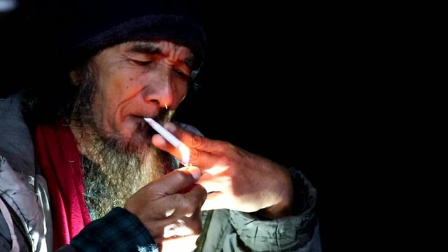 an old man with a beard and a thick jacket wearing a head covering while smoking at night