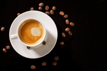 A small white cup of aromatic coffee with grains scattered around on a black background. Space for text.