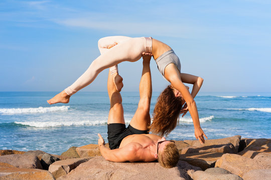 Fit young couple doing acro yoga at spa retreat on sea beach. Active woman balancing on partner feet, stretching at acroyoga pose. Healthy lifestyle. People outdoor sport activity on family vacation.