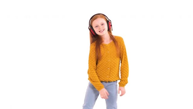 Little Caucasian girl with beautiful foxy hair and charming smile wearing wireless headphones and funnily dancing, relaxing and expressing positive emotions. Isolated, on white background