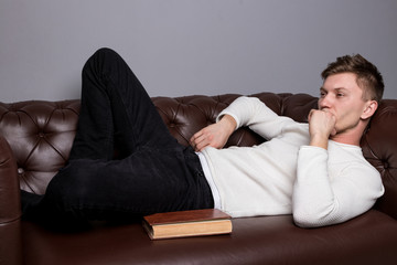 Young man is is lying on a leather sofa and reading a book