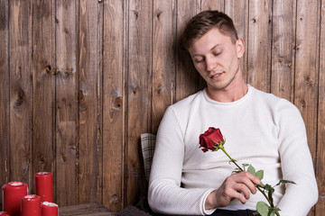 Young handsome man with a rose in his hands sitting near wood wall