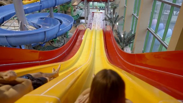 A woman and a man are in the water Park, they simultaneously roll down the water slides on their backs and plop into the water. The view from the back.
