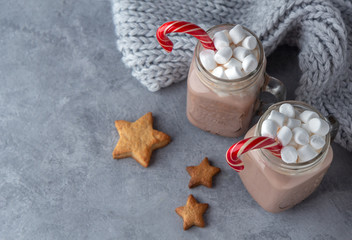 Hot cocoa with marshmallows and a sugar cane in glass mugs on a gray background with a knitted scarf.