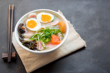 Asian style soup with udon noodles with steam and smoke, pork, boiled eggs, mushrooms and green onions close-up in a bowl on old table. horizontal top view from above